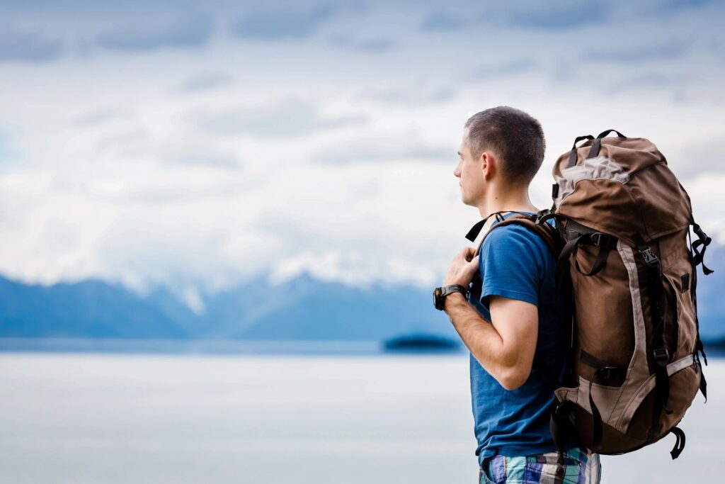 Man with backpack looking across lake at snow capped mountains. 